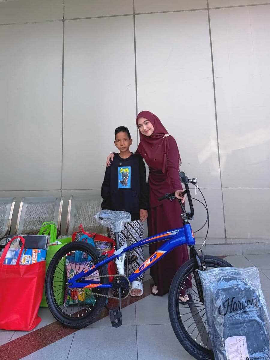 Seorang wanita berhijab berpose bersama seorang anak laki-laki di samping sepeda baru dan beberapa tas belanja berwarna cerah.