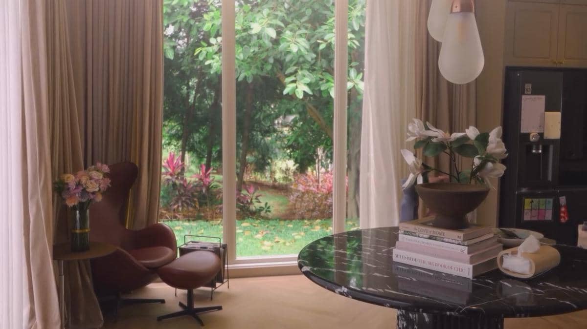 Ruang tamu rumah mewah dengan jendela besar menghadap taman hijau, kursi kulit cokelat, dan meja marmer hitam dengan vas bunga putih.