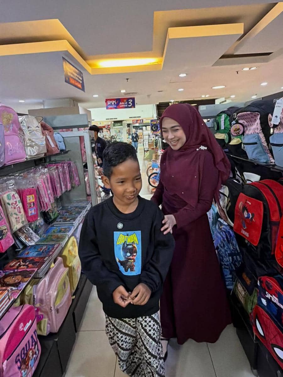 Seorang wanita berhijab berwarna marun tersenyum sambil menemani seorang anak laki-laki berbelanja di toko perlengkapan sekolah.