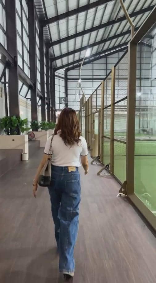 Seorang wanita berjalan di area dalam gedung lapangan padel dengan lantai kayu dan dinding kaca di sisi kanan.
