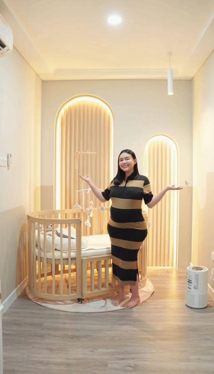 Seorang wanita berdiri tersenyum di kamar bayi dengan dekorasi minimalis bernuansa krem dan tempat tidur bayi kayu bundar.