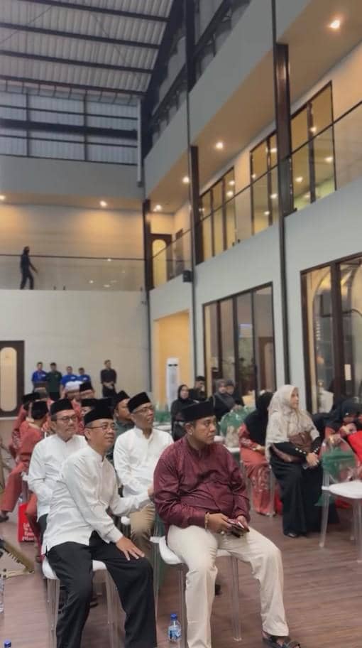 Sejumlah tamu duduk mengenakan busana formal dan peci hitam dalam acara syukuran di area dalam gedung berarsitektur modern bertingkat.