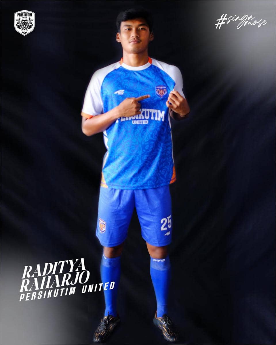 Seorang pemain sepak bola mengenakan seragam biru Persikutim United dengan latar belakang hitam, berdiri sambil menunjuk ke logo tim di dadanya.