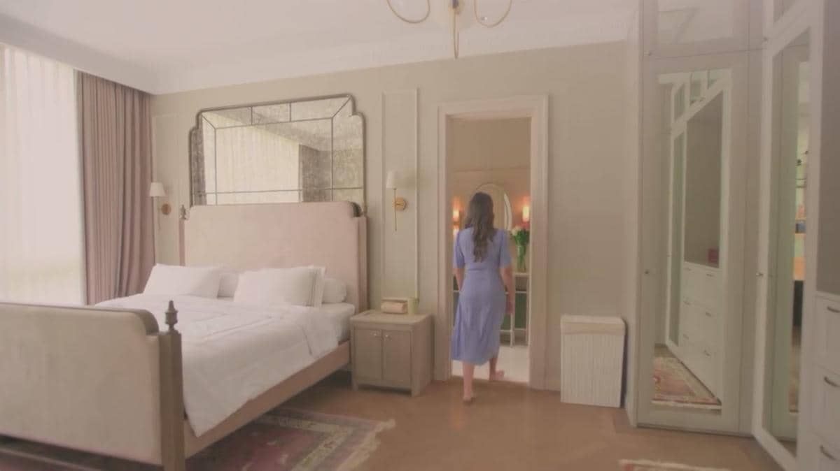 Seorang wanita mengenakan gaun biru berjalan di kamar tidur mewah dengan tempat tidur besar, cermin besar, dan pencahayaan lembut.