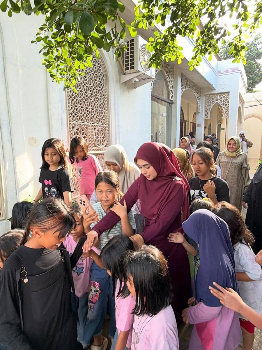 Seorang wanita berhijab berwarna marun berjalan bersama sekelompok anak perempuan di area luar bangunan bercorak islami.