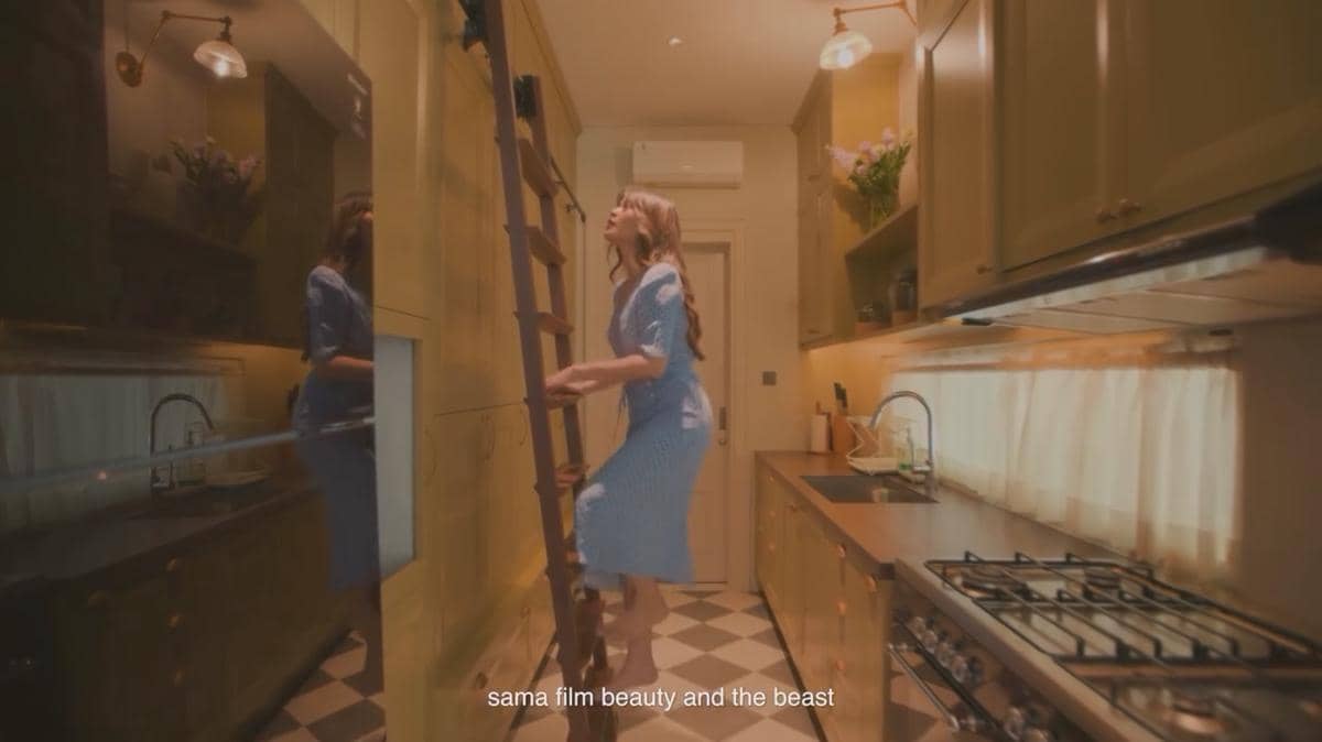 Seorang wanita mengenakan gaun biru sedang menaiki tangga kayu di dapur rumah mewah dengan desain interior elegan dan pencahayaan hangat.
