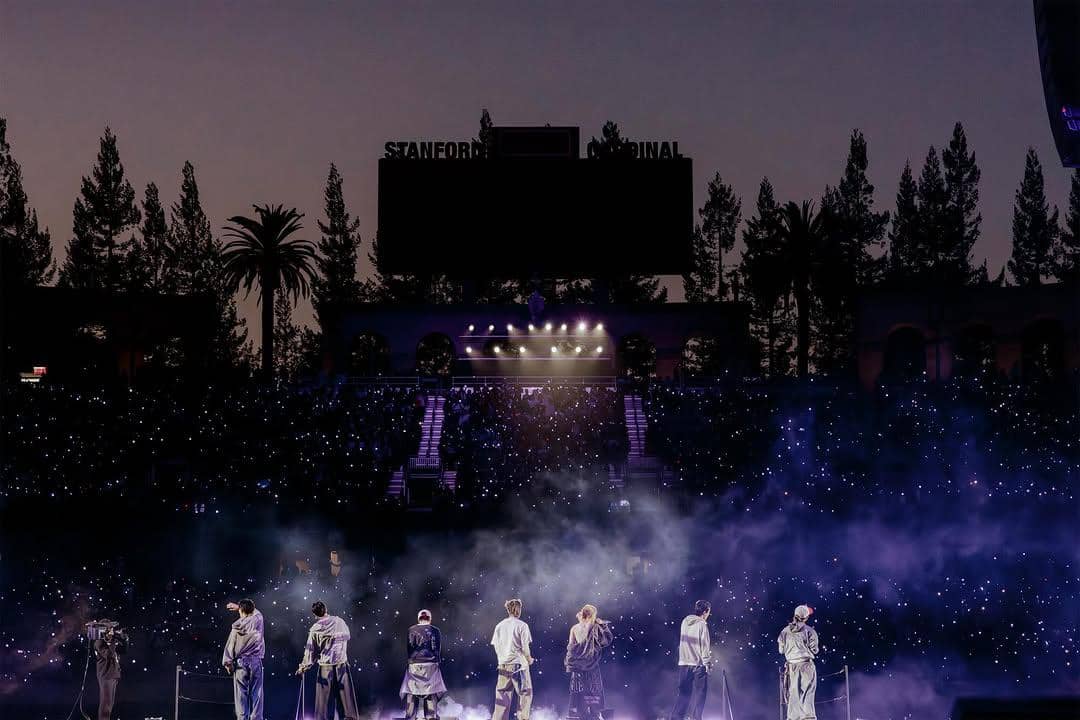Tujuh anggota grup musik tampil di atas panggung besar dengan pencahayaan ungu dan penonton memenuhi stadion saat malam hari.