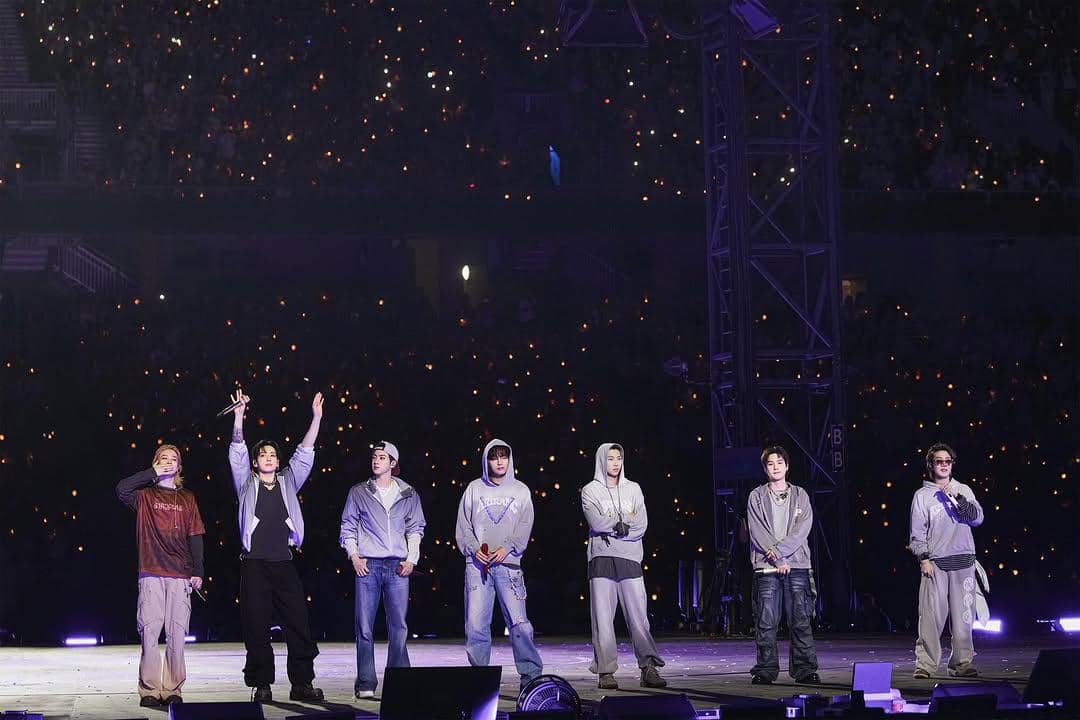Tujuh anggota grup musik berdiri di atas panggung besar dengan pencahayaan ungu dan lautan penonton memegang lampu di latar belakang.
