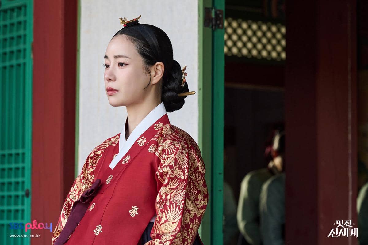 Seorang wanita mengenakan hanbok merah berhias bordir emas berdiri di depan bangunan tradisional Korea dalam adegan drama My Royal Nemesis.