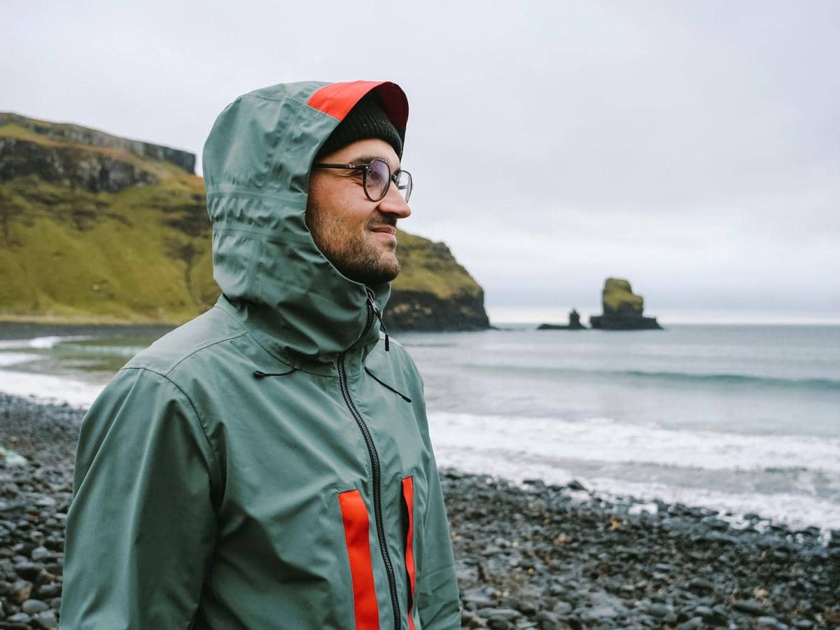 Seorang pria mengenakan jaket waterproof berwarna hijau dengan aksen merah berdiri di tepi pantai berbatu berlatar tebing dan laut.