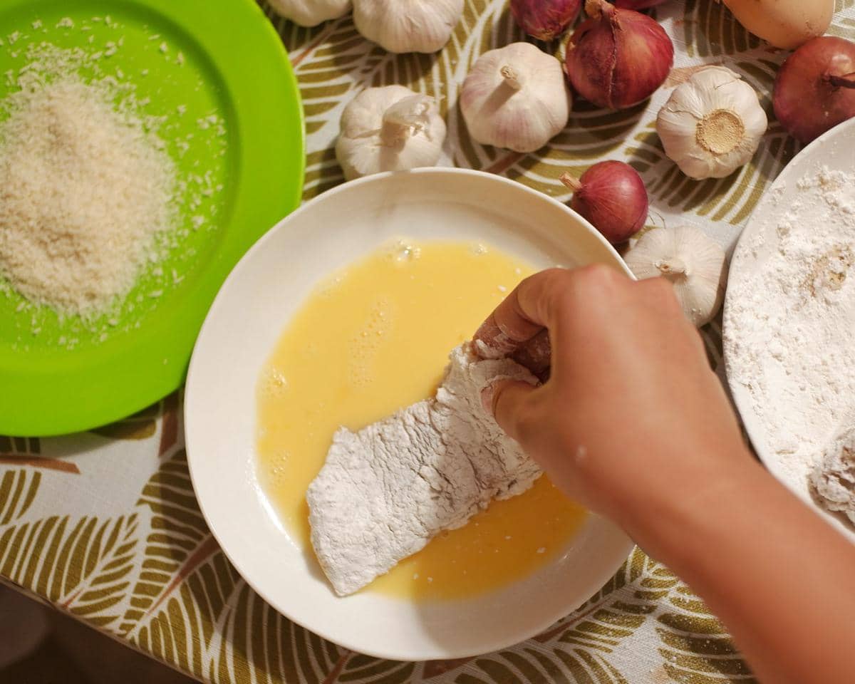Tangan seseorang mencelupkan potongan daging berbalut tepung ke dalam kocokan telur di atas meja dengan bawang putih dan bawang merah.
