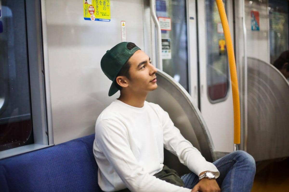 Seorang pria mengenakan topi hijau dan sweater putih duduk di kursi kereta, menatap ke arah jendela dengan ekspresi tenang.