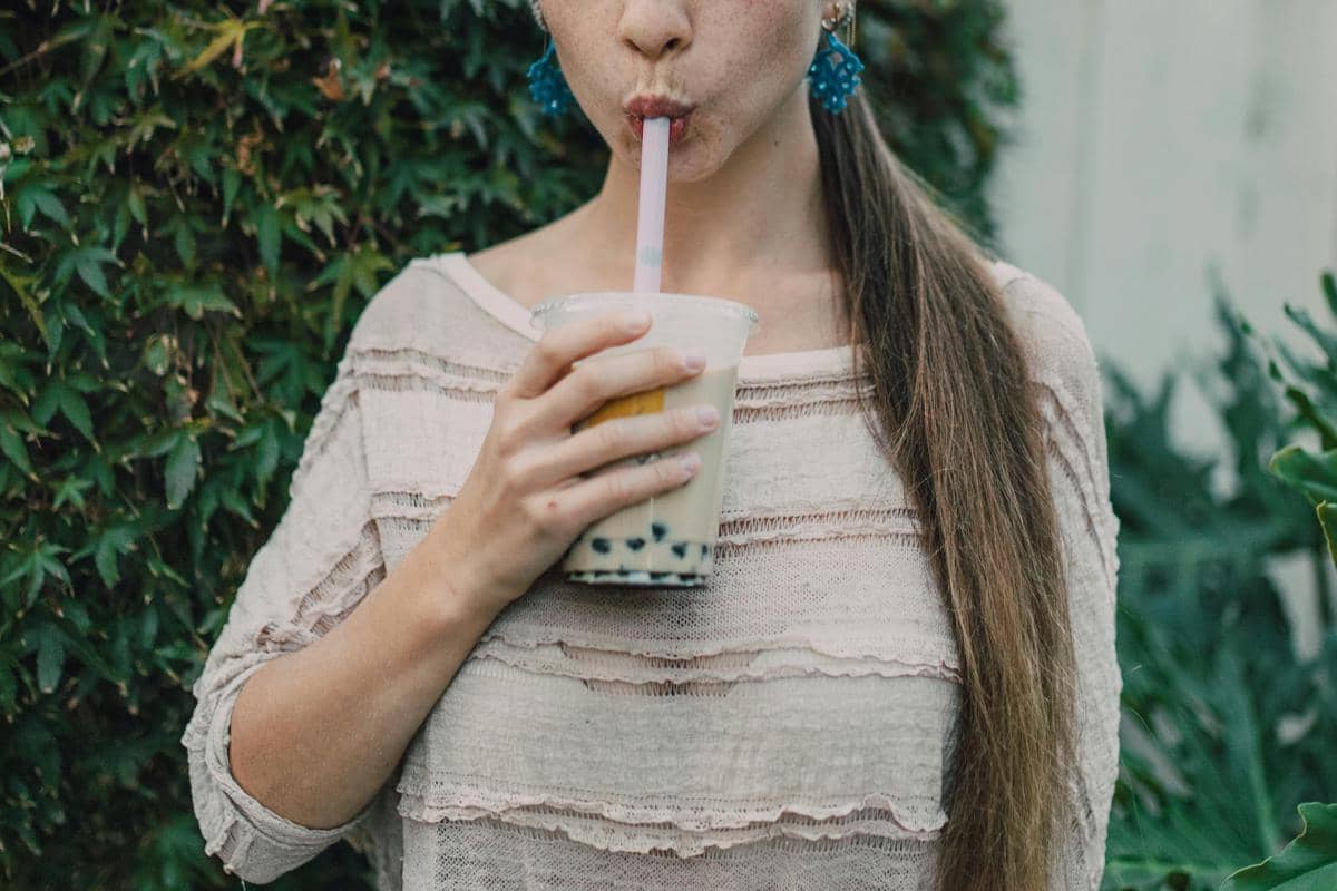 Seorang perempuan mengenakan blus krem sedang meminum segelas minuman manis boba dengan sedotan besar di luar ruangan.