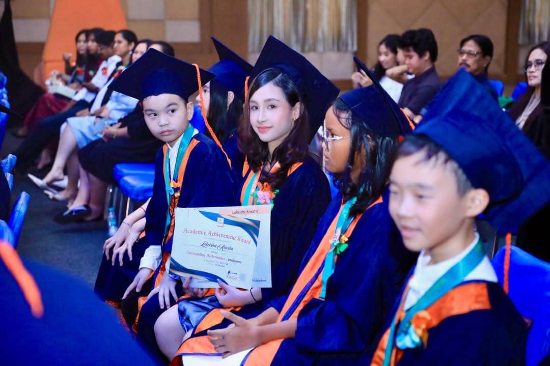 Anak-anak mengenakan toga biru tua dan topi wisuda duduk berbaris sambil memegang sertifikat kelulusan di acara wisuda sekolah dasar.