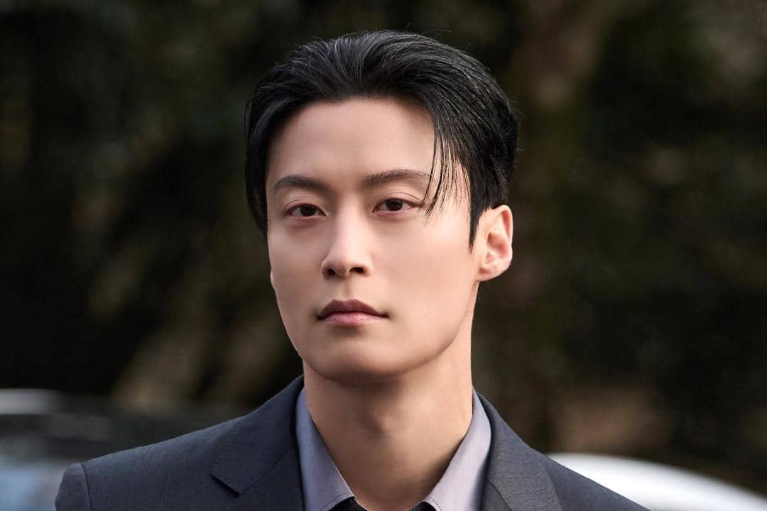 Seorang pria berambut hitam mengenakan setelan jas abu-abu tampak serius dalam cuplikan drama My Royal Nemesis.