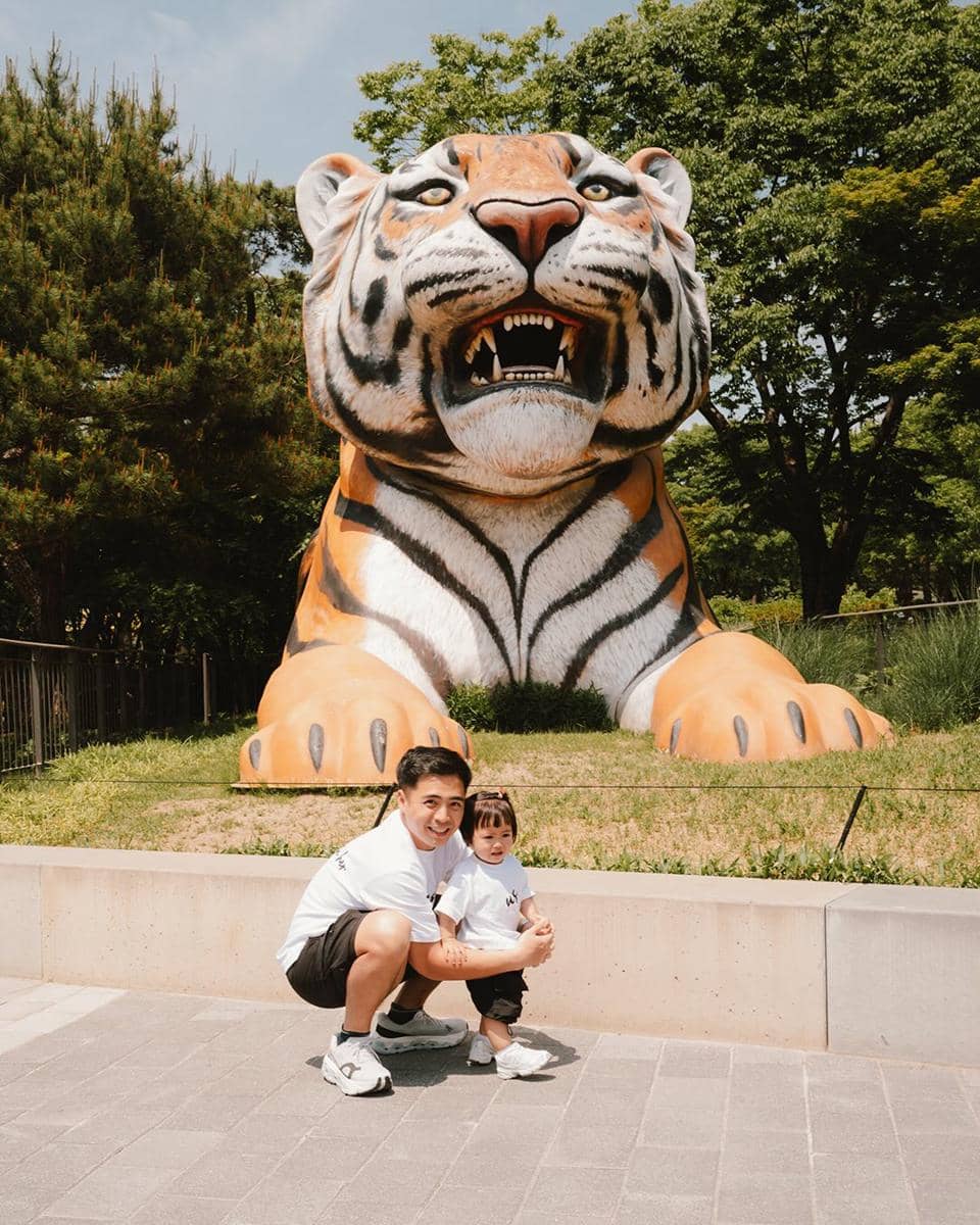 Seorang pria dan anak kecil berpose di depan patung harimau besar di area terbuka dengan pepohonan hijau di Gangnam.