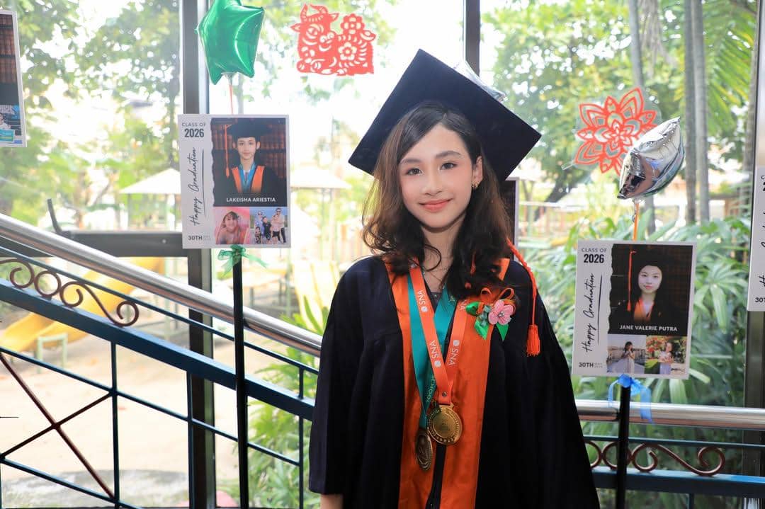 Seorang anak perempuan mengenakan toga wisuda lengkap dengan medali dan selempang berwarna oranye saat acara kelulusan sekolah dasar.