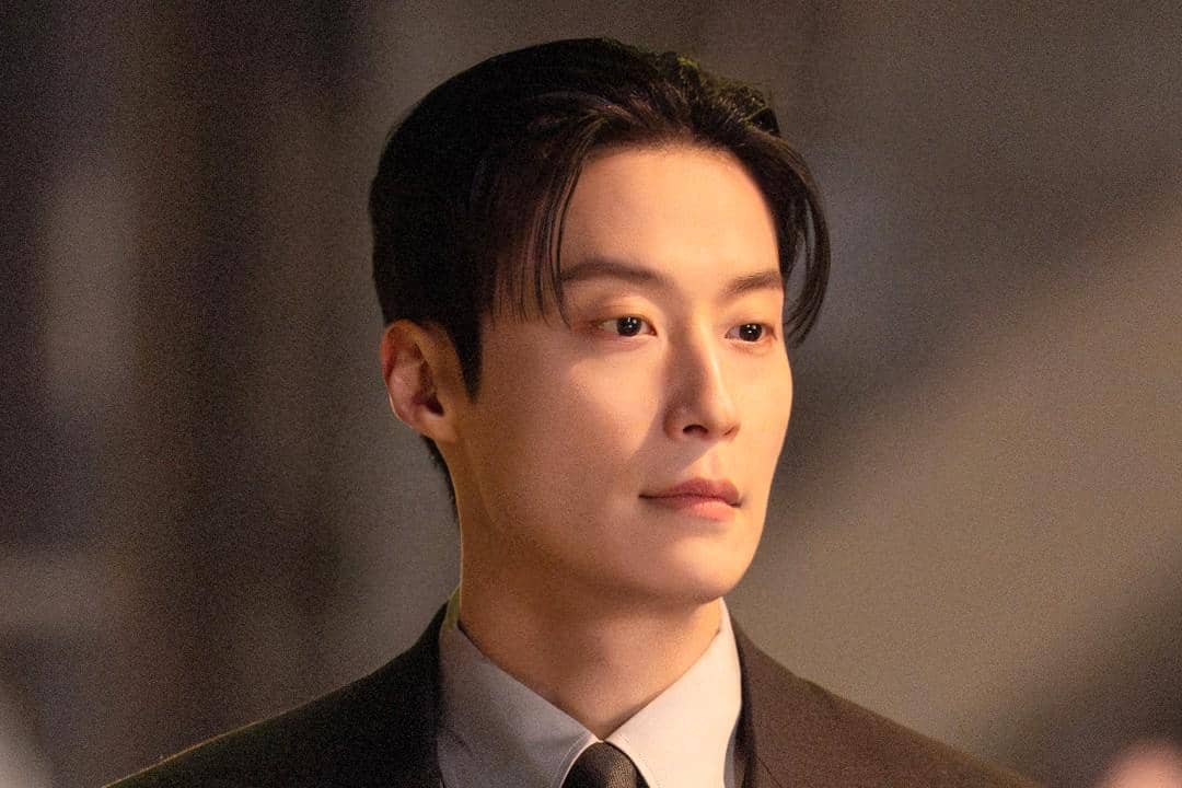 Seorang pria berpenampilan rapi mengenakan setelan jas dan dasi dalam adegan drama My Royal Nemesis dengan pencahayaan hangat.