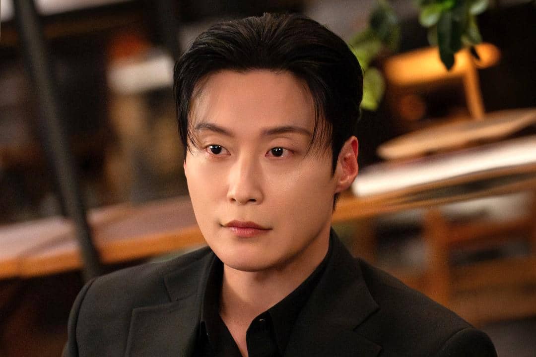 Seorang pria berpenampilan rapi mengenakan setelan jas hitam dengan ekspresi serius dalam adegan drama My Royal Nemesis.
