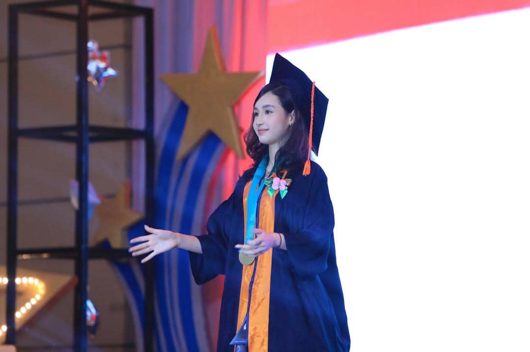 Seorang anak mengenakan toga wisuda berwarna biru tua dengan selempang oranye dan biru di atas panggung acara kelulusan sekolah dasar.