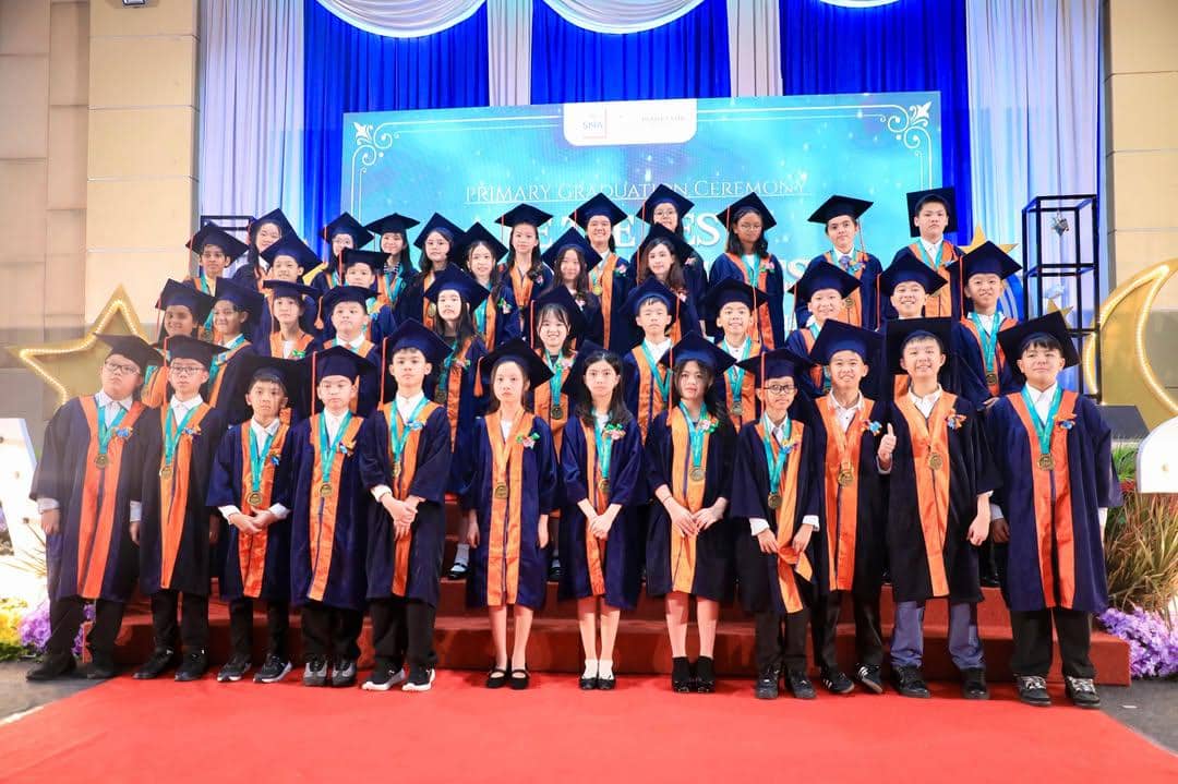 Sekelompok siswa sekolah dasar mengenakan toga biru dan oranye berpose di atas panggung saat acara kelulusan dengan latar biru berhias bintang.