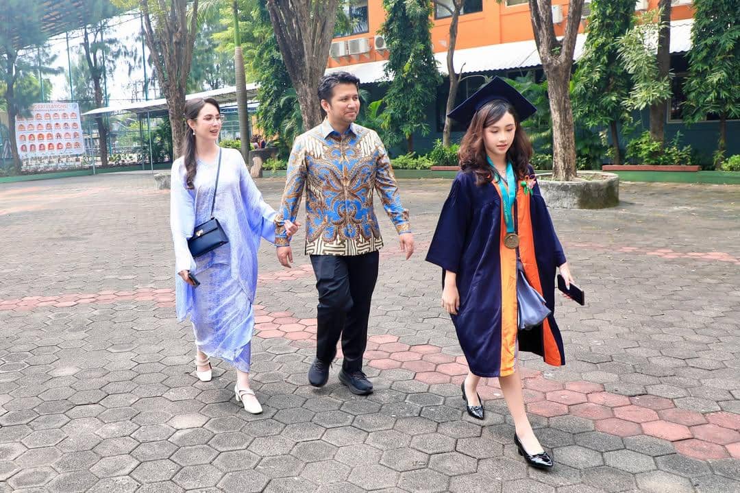 Seorang anak mengenakan toga wisuda berjalan bersama dua orang dewasa di area sekolah dengan latar pepohonan dan gedung oranye.