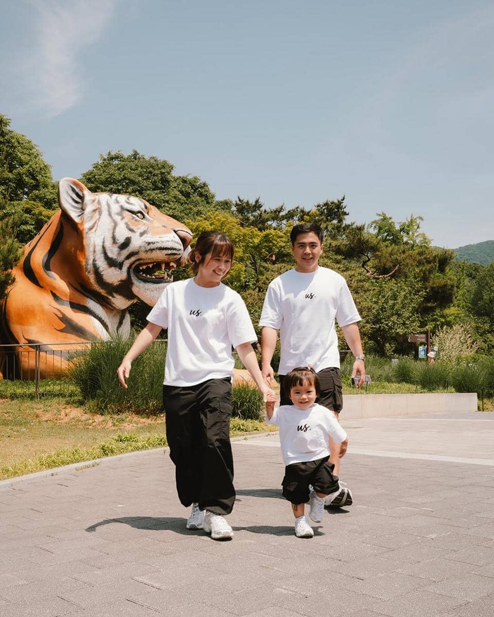 Sebuah keluarga berjalan bersama di area terbuka Gangnam, mengenakan kaus putih dan celana hitam dengan latar patung harimau besar.