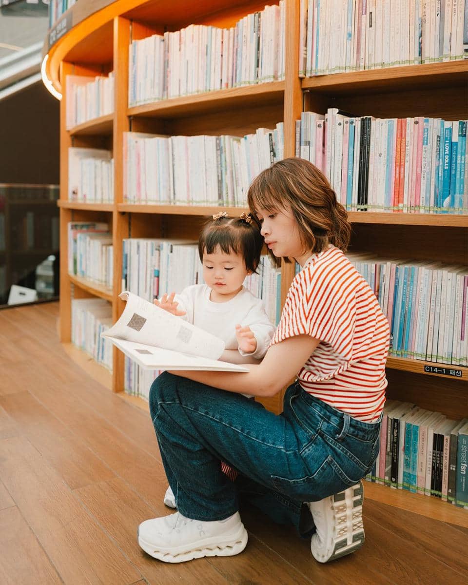 Seorang wanita mengenakan kaus bergaris merah putih sedang membaca buku bersama anak kecil di perpustakaan dengan rak buku tinggi.