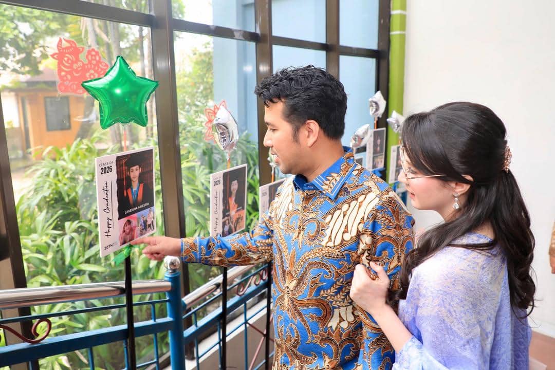 Sepasang orang tua mengenakan busana batik dan kebaya melihat papan foto kelulusan anak di acara wisuda sekolah dasar.