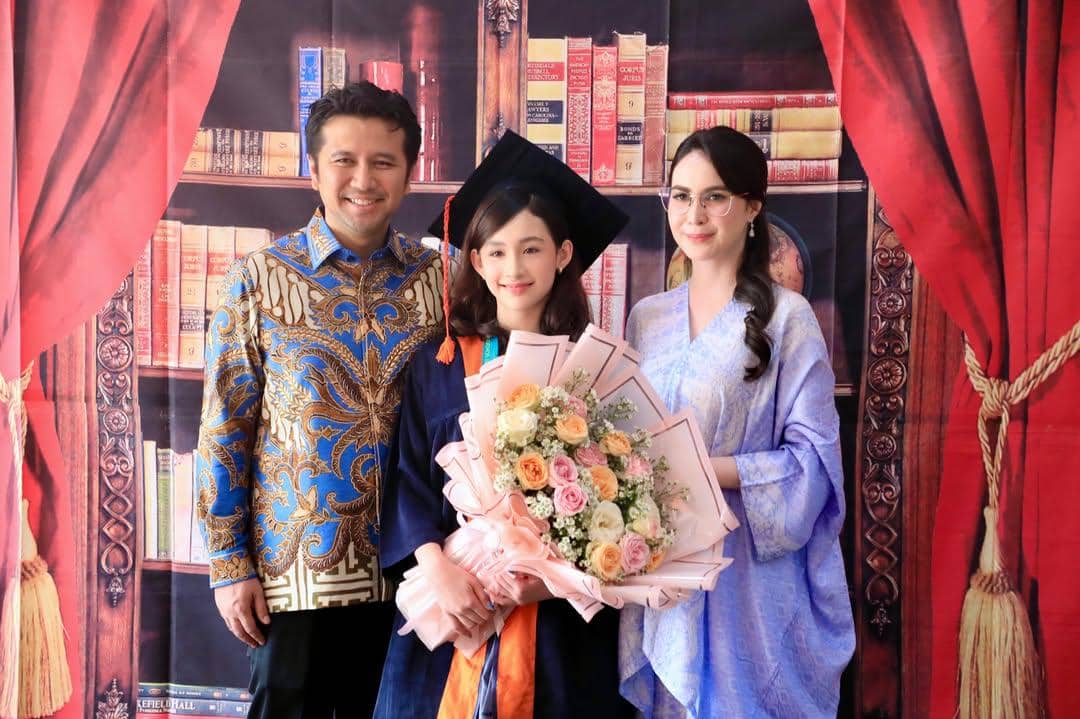 Seorang anak mengenakan toga wisuda SD memegang buket bunga berpose bersama dua orang dewasa di depan latar rak buku dan tirai merah.