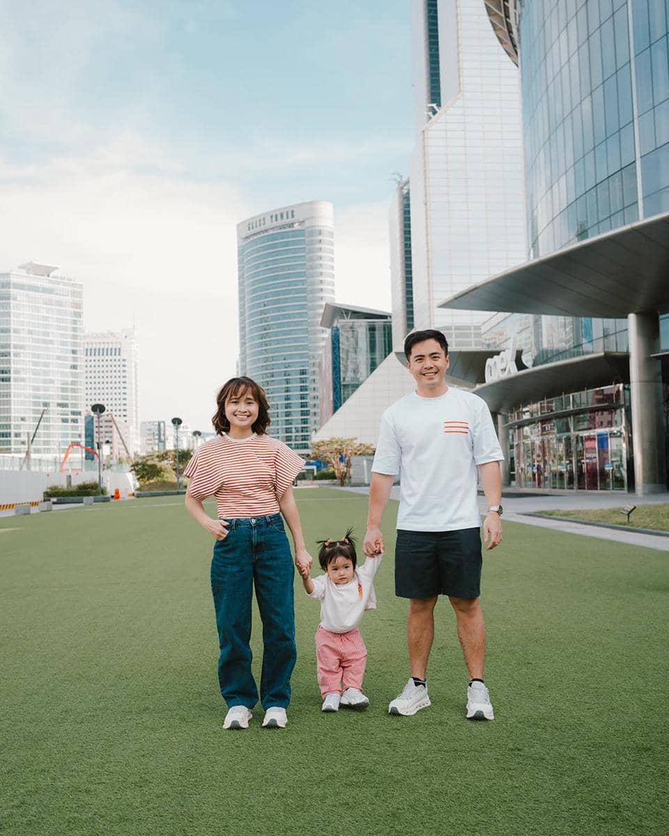 Sebuah keluarga terdiri dari dua orang dewasa dan seorang anak kecil berpose sambil bergandengan tangan di area hijau Gangnam dengan latar gedung tinggi modern.