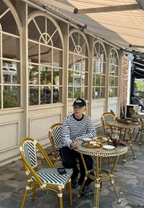 Seorang pria mengenakan kaus garis-garis hitam putih dan topi hitam duduk di kafe luar ruangan dengan meja berisi makanan dan minuman.