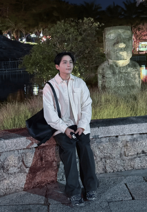 Seorang pria berambut hitam mengenakan kemeja putih dan celana gelap duduk di tepi taman dengan patung batu besar di belakangnya pada malam hari.