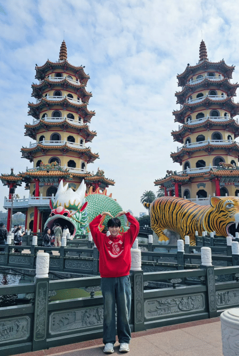 Seseorang berpose mengenakan sweater merah di depan Menara Naga dan Harimau di Danau Lotus, Kaohsiung, dengan langit cerah di latar belakang.