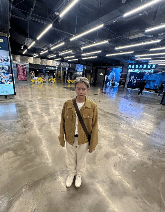 Seseorang berdiri di area bioskop modern dengan pencahayaan terang, mengenakan jaket cokelat muda, celana putih, dan tas selempang.