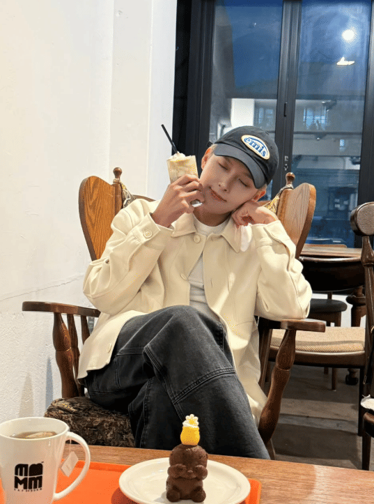 Seorang pria mengenakan jaket krem dan topi biru duduk santai di kafe sambil memegang minuman dengan mata terpejam.