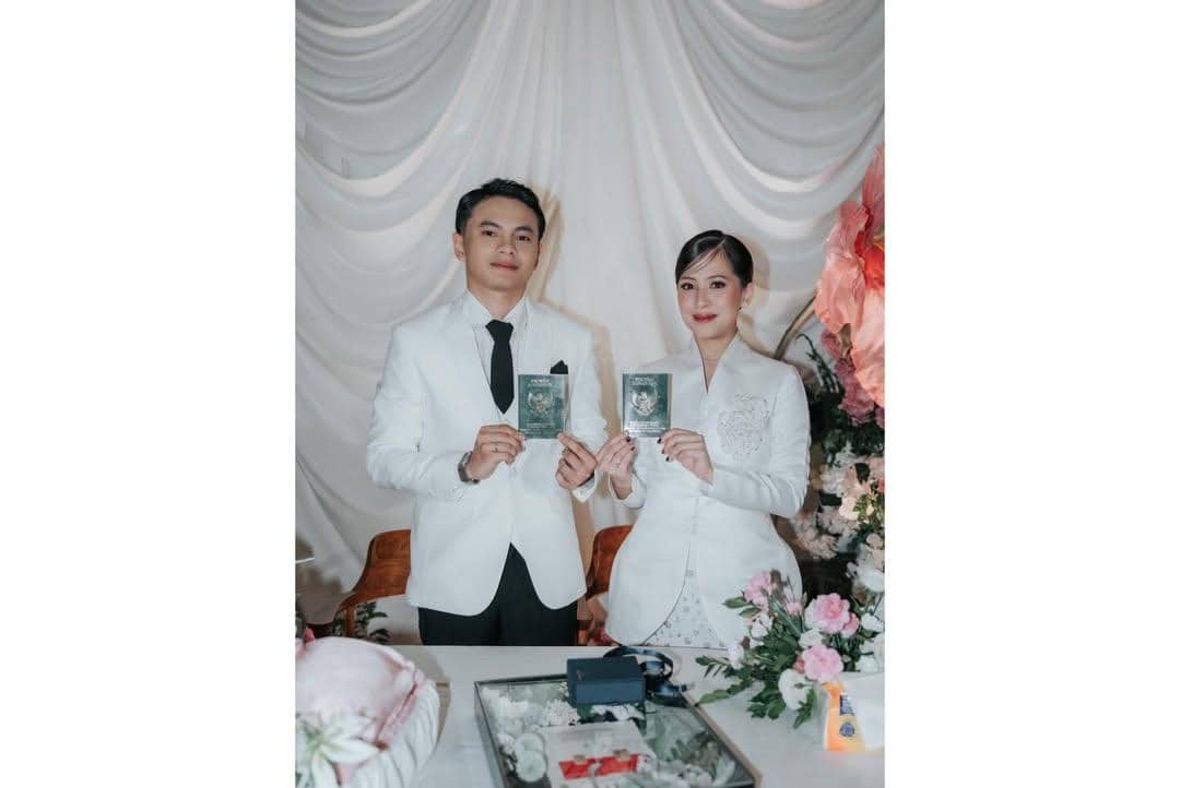 Sepasang pengantin mengenakan busana putih memegang buku nikah di depan dekorasi kain putih dan bunga berwarna lembut.