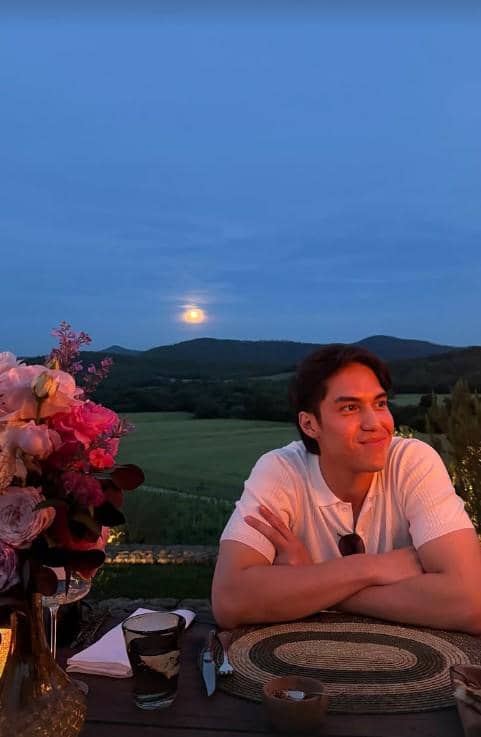Seorang pria duduk di meja makan luar ruangan saat senja dengan latar perbukitan hijau dan bulan purnama di Italia.