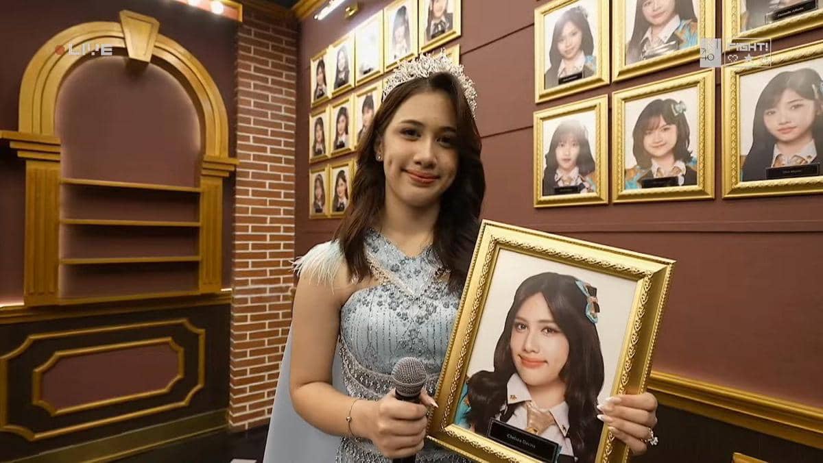 Seorang anggota JKT48 mengenakan gaun biru muda memegang mikrofon dan bingkai foto dirinya saat acara kelulusan di studio.