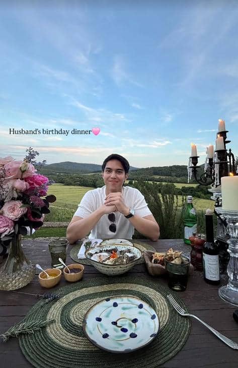 Seorang pria duduk di meja makan luar ruangan dengan pemandangan perbukitan Italia, dihiasi bunga, lilin, dan hidangan ulang tahun.