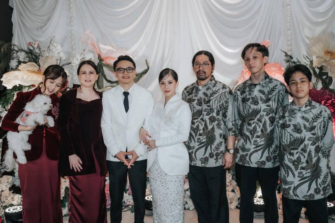 Pasangan pengantin berbusana putih berpose bersama keluarga dalam acara pernikahan dengan latar dekorasi bunga dan tirai putih.