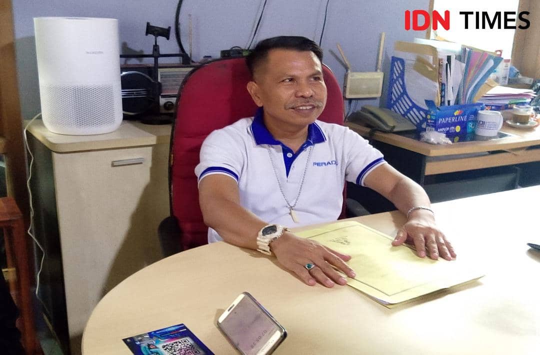 Seorang pria mengenakan kaus berkerah putih duduk di kursi merah di belakang meja kantor dengan berkas dan ponsel di depannya.