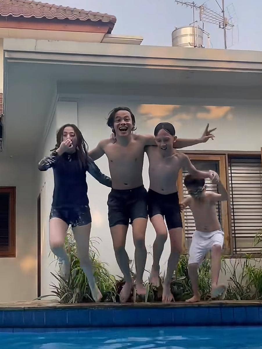 Empat orang, dua pria dan dua anak, melompat bersama ke kolam renang di halaman rumah dengan ekspresi gembira.