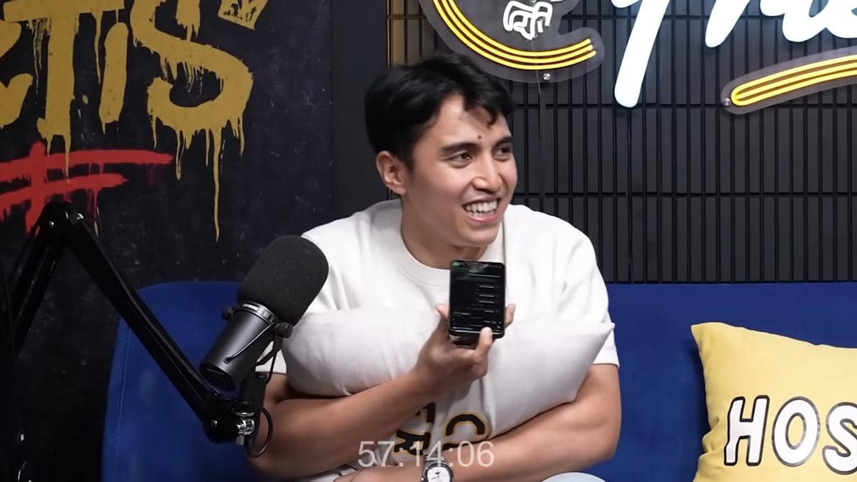 Seorang pria tersenyum sambil memegang ponsel dan bantal di studio podcast dengan mikrofon di depannya dan latar bertuliskan Friends.