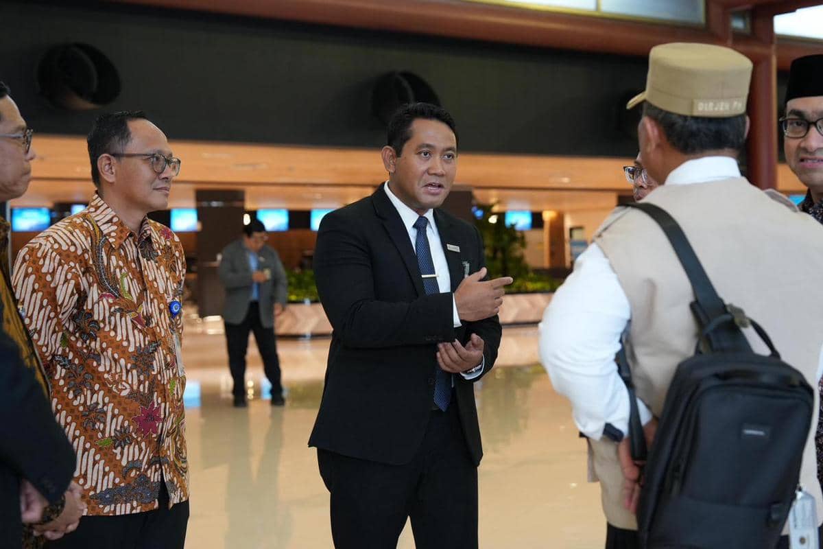 Seorang pria berpakaian jas hitam berbicara dengan beberapa orang di area bandara dengan suasana profesional dan formal.