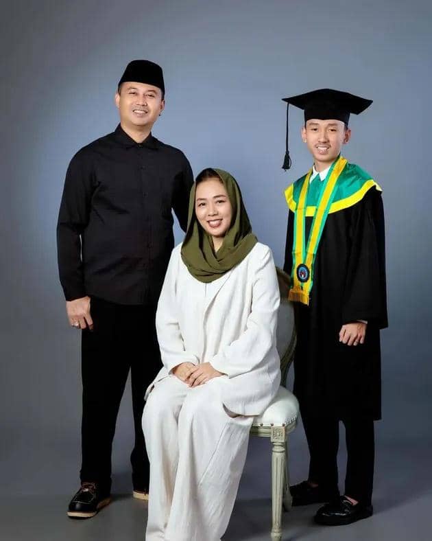 Seorang lulusan mengenakan toga hitam berpose bersama dua orang dewasa, satu duduk mengenakan hijab hijau dan satu berdiri mengenakan pakaian hitam.