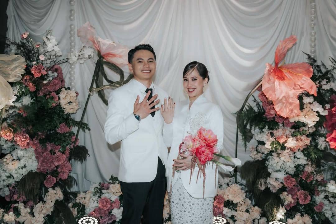 Sepasang pengantin mengenakan busana putih berpose sambil memperlihatkan cincin pernikahan di depan dekorasi bunga berwarna lembut.