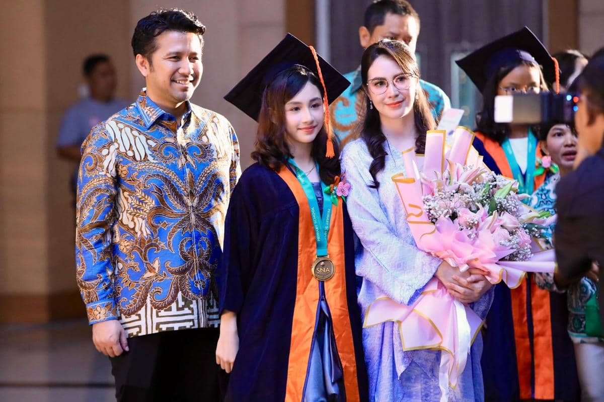 Seorang siswi mengenakan toga wisuda berfoto bersama orang tuanya yang tersenyum dan membawa buket bunga besar di acara kelulusan.