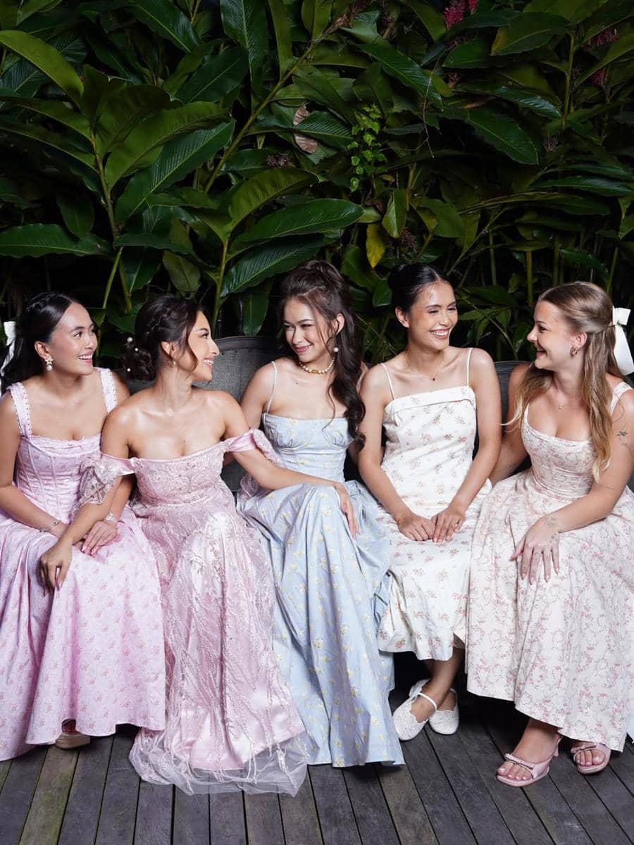 Lima wanita mengenakan gaun pastel duduk berdampingan sambil tersenyum di acara bridal lunch dengan latar daun hijau tropis.
