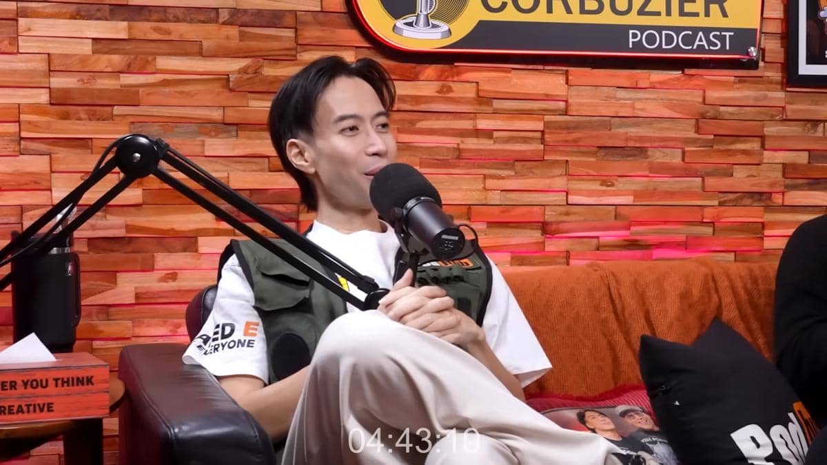 Seorang pria duduk santai di sofa studio podcast dengan mikrofon di depannya, mengenakan kaus putih dan rompi hijau.